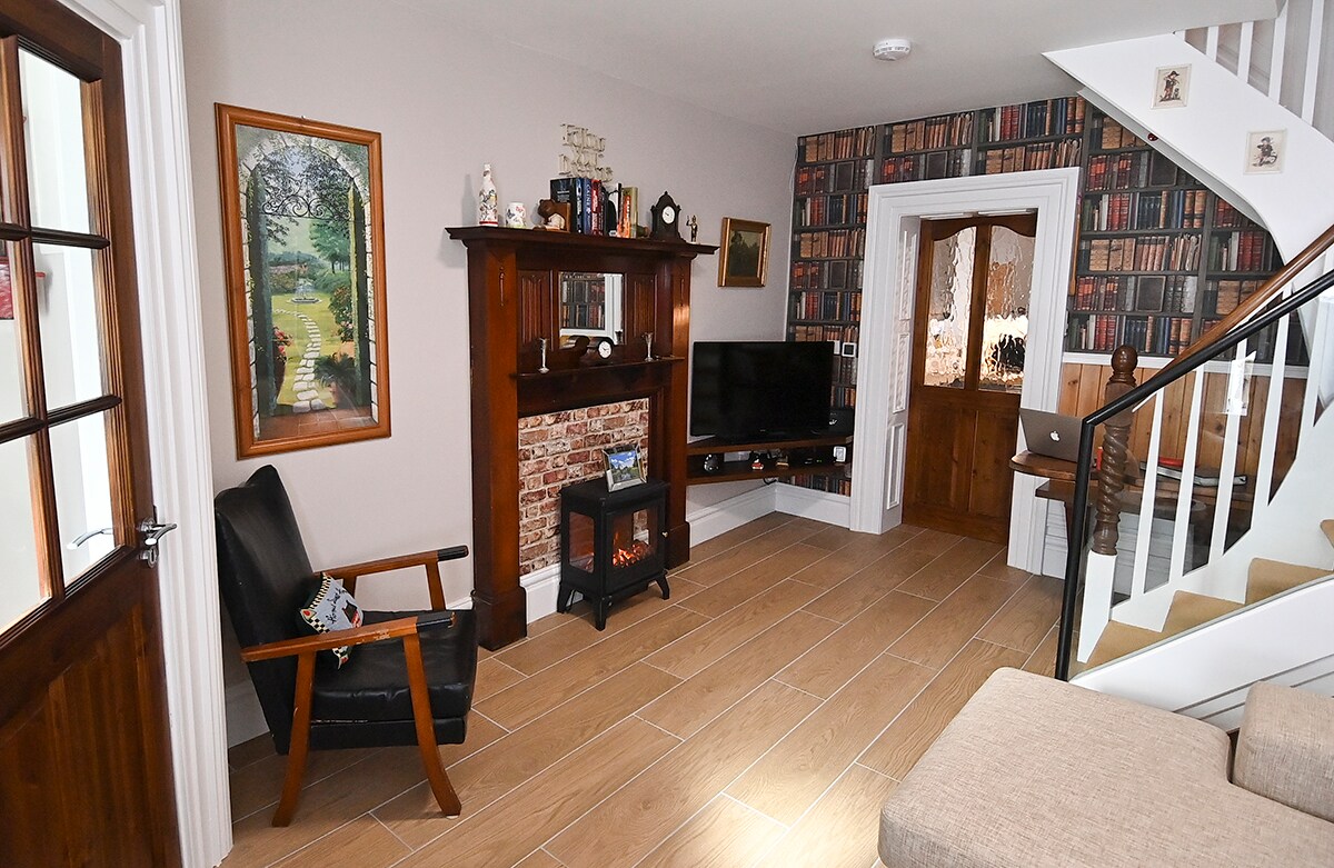 A cozy living area features an antique fireplace with an electric stove, surrounded by wooden shelving and decor. A comfortable black chair adds seating, while a smart TV is positioned for viewing. Warm lighting enhances the inviting space with patterned wallpaper in the background.