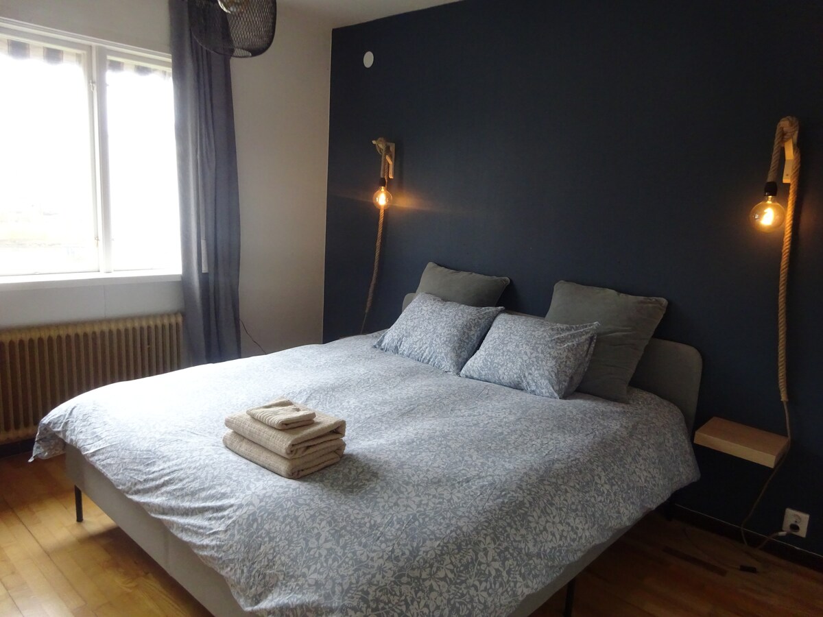 A spacious bed is positioned against a dark blue wall, complemented by two bedside lights. Light-colored bedding features a floral pattern. A set of neatly stacked towels rests on the bed, and natural light fills the room from the window, which is partially covered by sheer curtains.