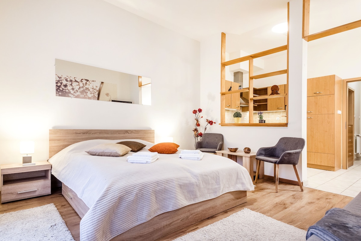 A spacious bedroom features a comfortable bed adorned with neatly arranged linens and pillows. Soft light from bedside lamps illuminates the room, accentuated by a large mirror above the bed. A small sitting area with two chairs and a table complements the inviting atmosphere.