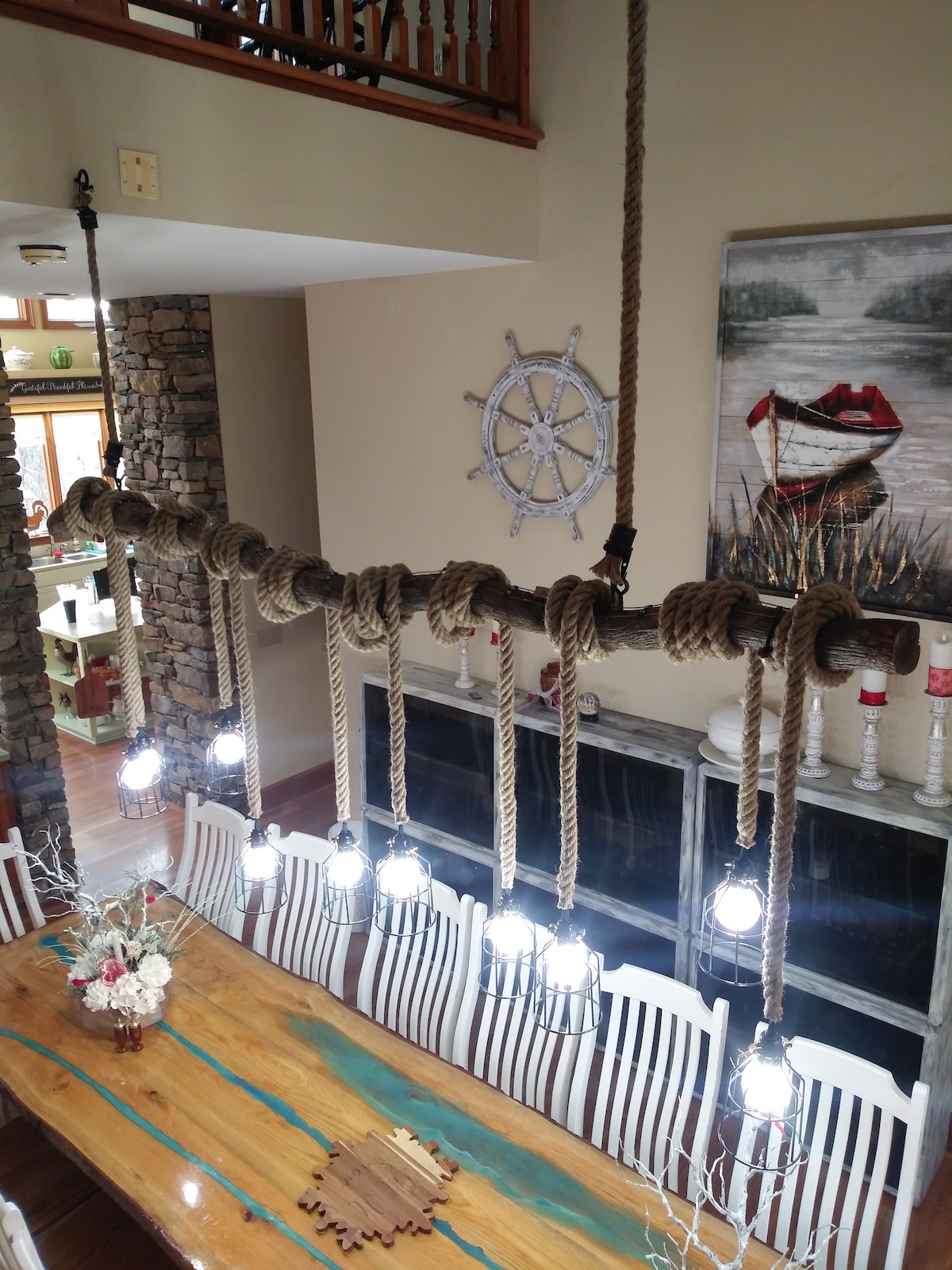 A large dining area is showcased with a long wooden table surrounded by white chairs. Above, a unique lighting fixture is suspended, featuring multiple lanterns intertwined with thick ropes. A decorative piece, resembling a ship's wheel, is mounted on the wall, complementing the rustic decor.
