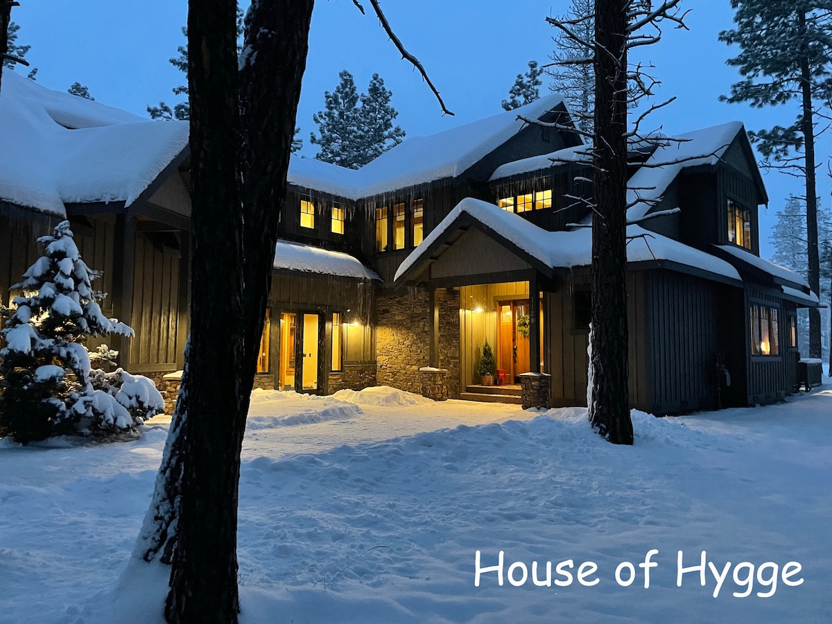 A spacious vacation home is surrounded by snow-covered trees, showcasing a welcoming exterior. Warm light glows from the windows, highlighting the charming architectural features and inviting entryway. Snow blankets the ground, adding a serene atmosphere to the picturesque setting.