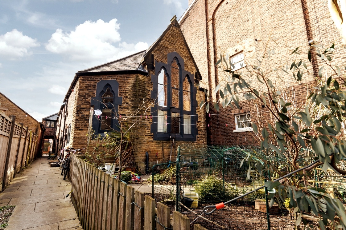 The exterior of a charming mews house is displayed, featuring large arched windows that provide a unique architectural detail. A garden area can be seen, with greenery and outdoor seating alongside a pathway leading to the entrance.