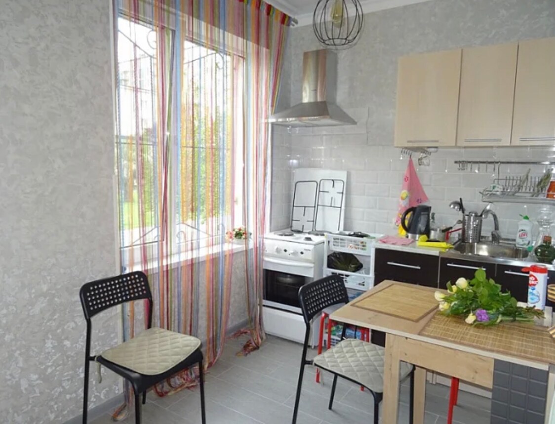 A functional kitchen area is showcased, featuring a cooking range, sink, and shelving. Two chairs are positioned at a small wooden table. Colorful curtains frame the window, allowing natural light to illuminate the space, highlighting the neutral tones of the walls and cabinets.