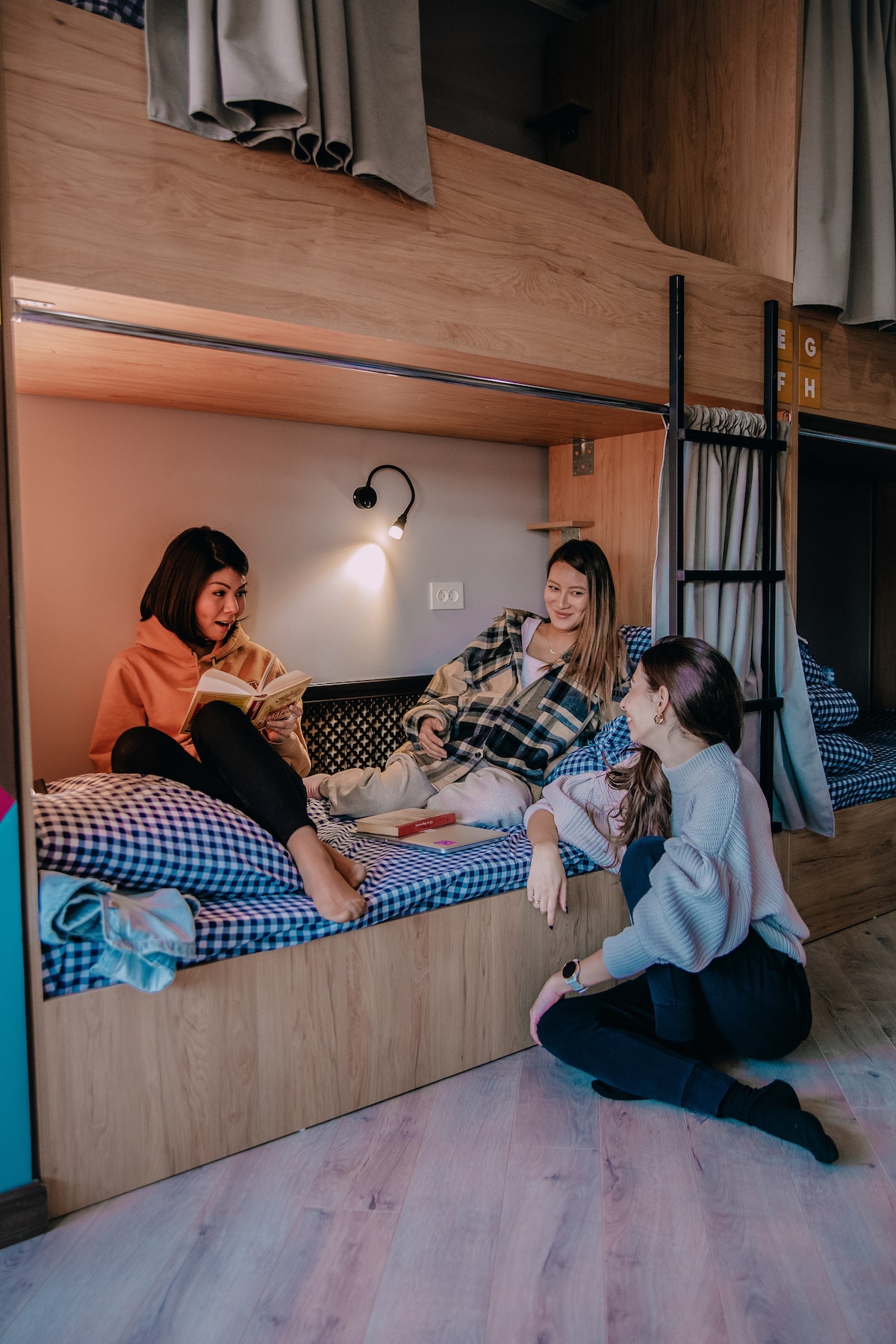 A cozy shared sleeping area is depicted, featuring wooden bunk beds with checkered bedding. Two individuals are seated on the lower bunk, engaged in conversation, while a third person is crouched beside them. Soft lighting is provided by a wall-mounted lamp, illuminating the warm tones of the wood.