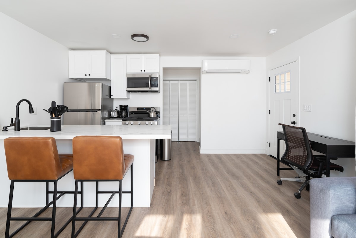 A modern kitchen area features a white countertop and two leather barstools. Stainless steel appliances include a fridge, stove, and microwave. A workspace with a desk and chair sits in the corner, while an air conditioning unit is mounted on the wall. Natural light brightens the open layout.