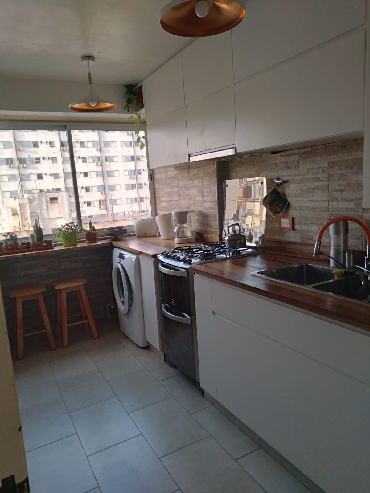 A modern kitchen is depicted, featuring sleek cabinetry and a spacious countertop. Large windows provide ample natural light and an urban view. Included are a washing machine, stove, sink, and a small breakfast area with two wooden stools. Potted plants add a touch of greenery to the space.