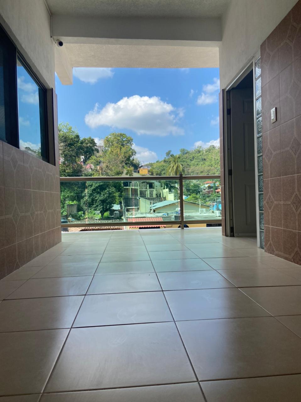 A spacious hallway leads to an outdoor balcony, featuring large windows that reveal a view of lush greenery and a clear blue sky. The tiled floor adds a modern touch, while the open door creates a welcoming entrance to the tranquil surroundings.