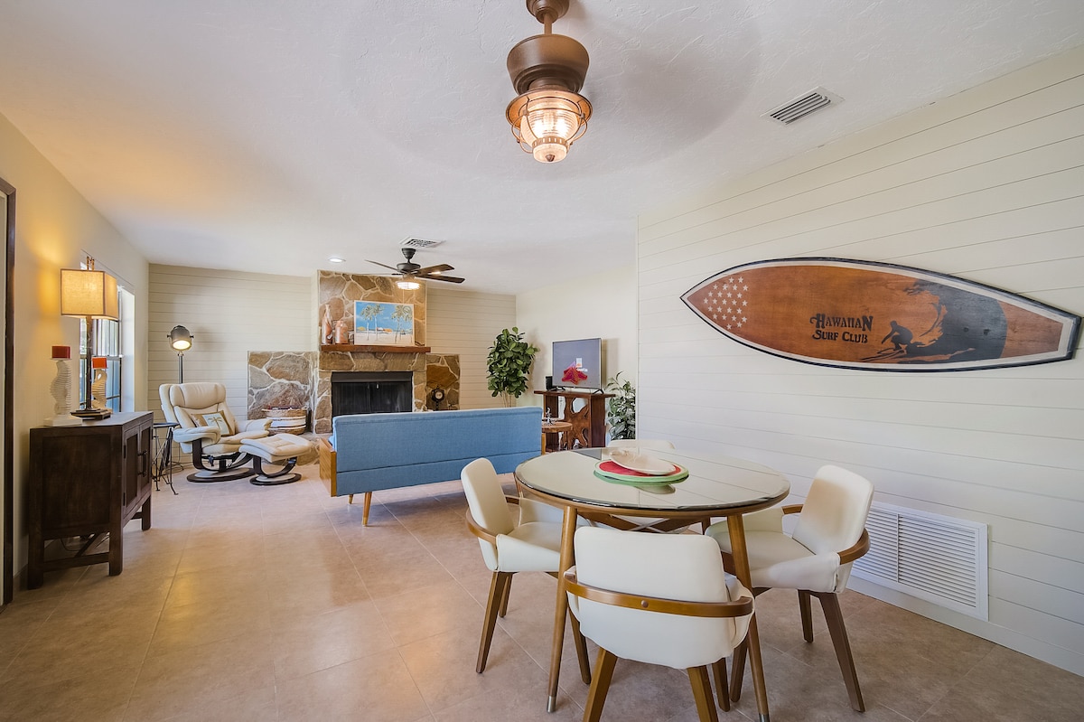 A bright living space features a combination of a cozy seating area with a blue sofa and a rustic stone fireplace. A round dining table surrounded by four light-colored chairs is positioned nearby. Decorative elements include a surfboard mounted on the wall and indoor plants.