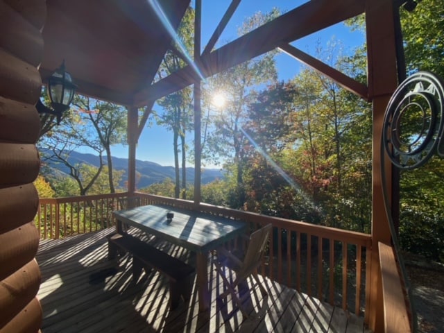 An outdoor deck features a spacious picnic table set against a backdrop of vibrant trees and mountains. Sunshine filters through the space, casting shadows on the wood deck. The natural scenery creates a serene and inviting environment.