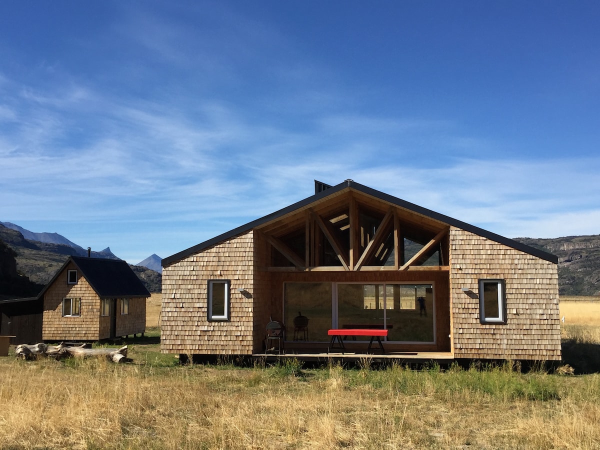 A modern wooden house features a large front deck with a red table and chairs. The structure is surrounded by open fields and mountains in the background. Large windows provide ample natural light and offer expansive views of the landscape.