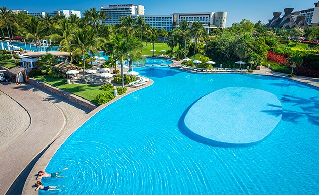 A large outdoor pool adorned with palm trees features sun loungers along its edges. The water exhibits a clear, inviting blue hue. Lush green lawns and tropical landscaping extend around the pool area, enhancing the serene atmosphere of the resort.