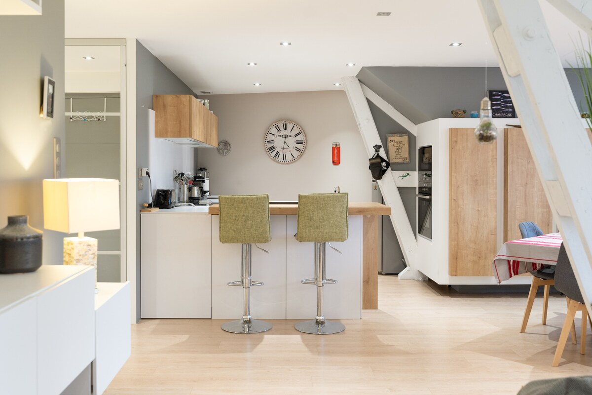 A modern kitchen area features sleek white cabinetry and a central island with two high stools. A clock hangs prominently on the wall. Various kitchen appliances are arranged neatly, and natural light illuminates the space through large windows.