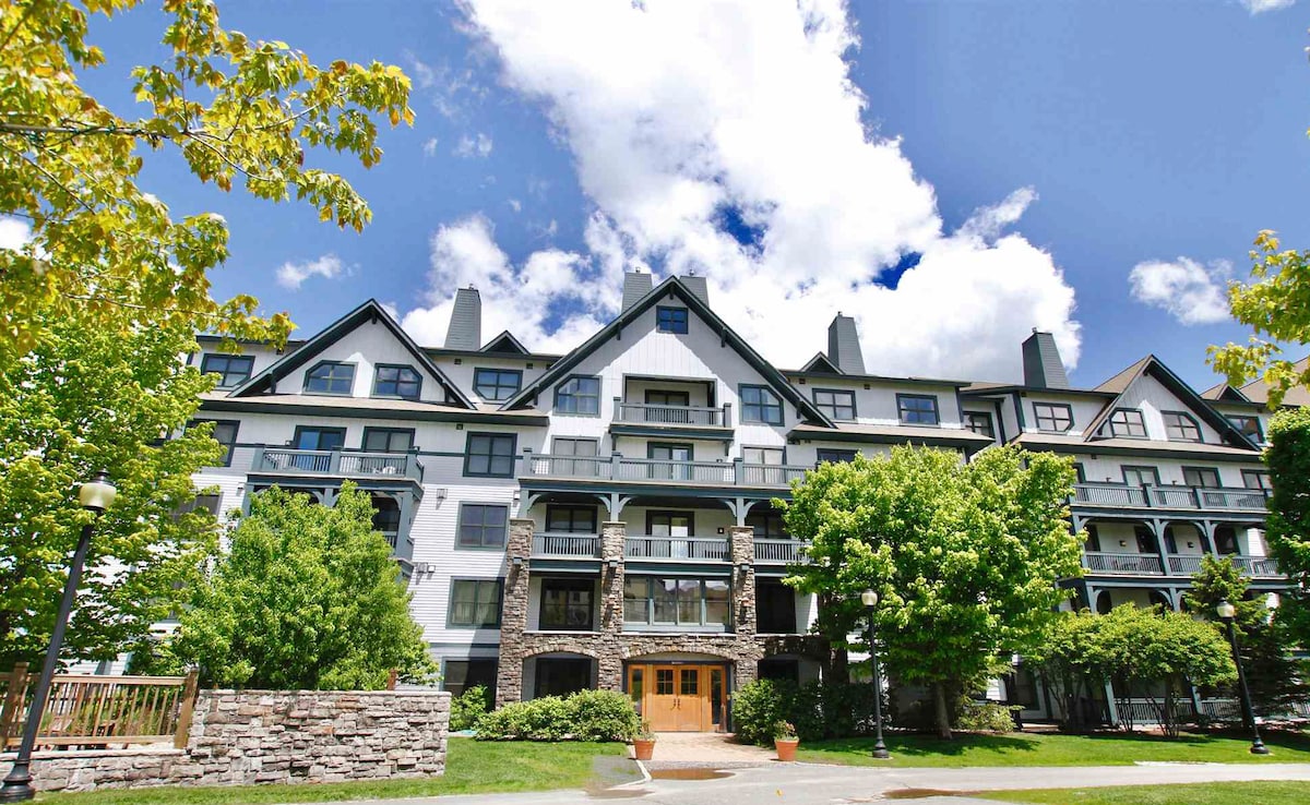 The exterior of the lodge is displayed against a bright blue sky with fluffy white clouds. Stone and wood accents characterize the building's design, featuring balconies that overlook landscaped green areas and trees.