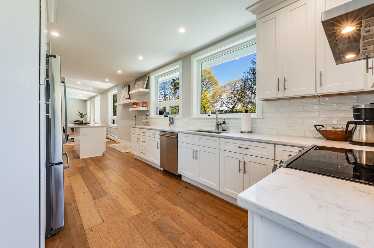 A modern kitchen features white cabinetry and stainless steel appliances. Large windows allow natural light to fill the space, highlighting the wooden flooring. A spacious island is positioned in the center, providing additional counter space for cooking and preparation.