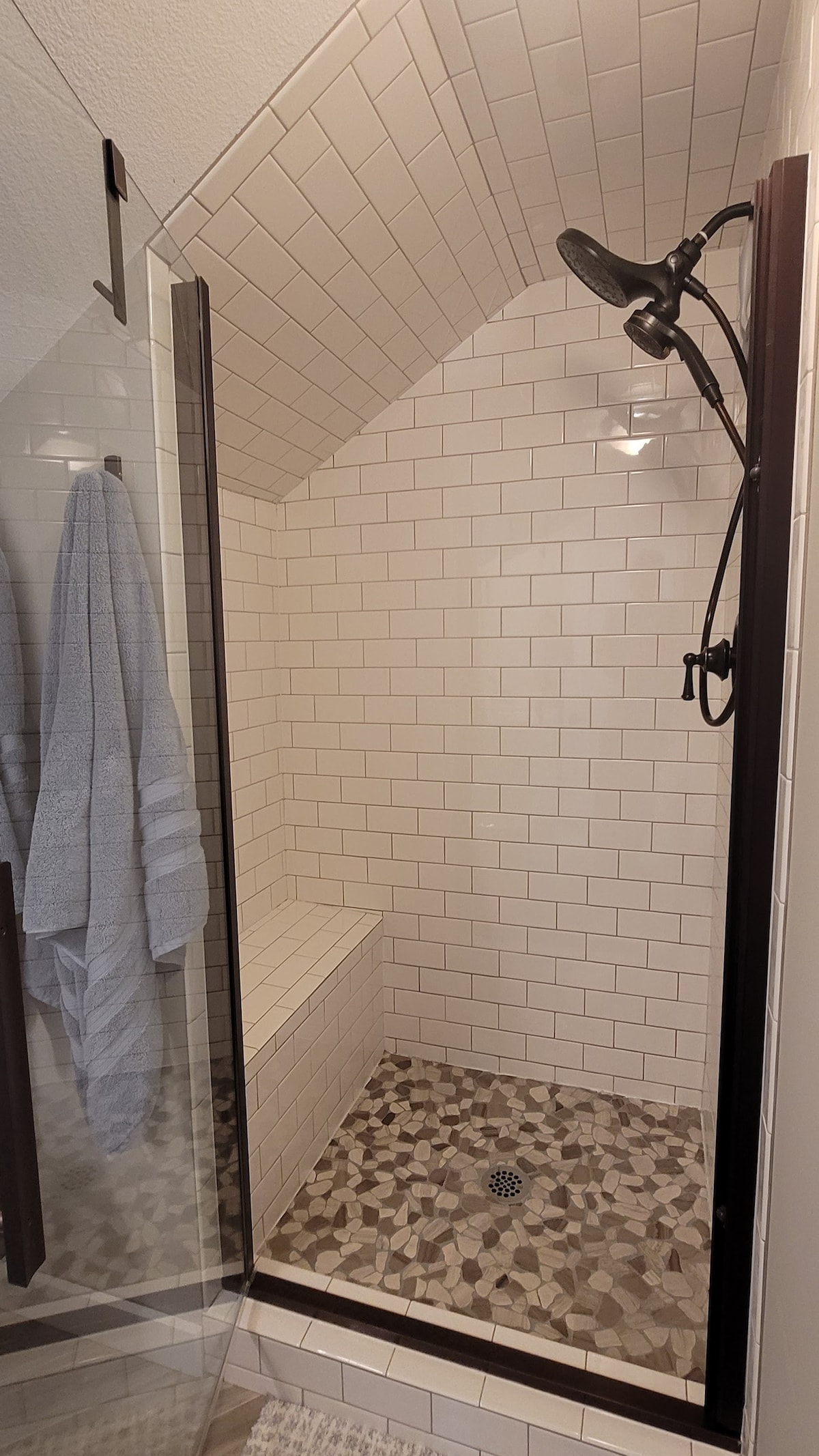 A walk-in shower features white subway tiles with a sloped ceiling, showcasing a built-in bench. The floor is adorned with small, decorative stones. A rain showerhead is mounted above, and a grey towel hangs on a hook nearby.