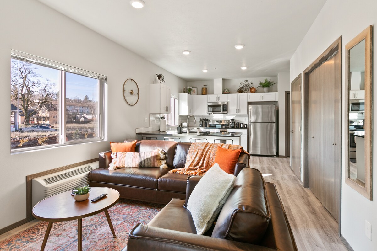 A modern living area features a leather sectional couch adorned with decorative pillows and a cozy throw. Natural light fills the space through a large window, complementing the open kitchen with stainless steel appliances and wood-style flooring.