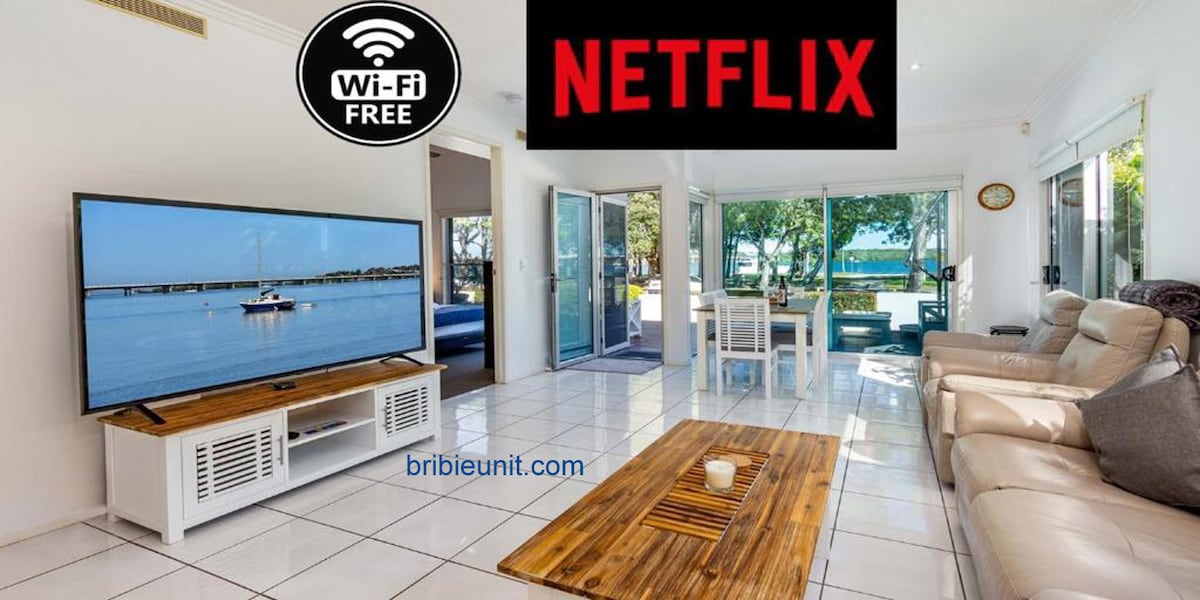 A bright living area is shown, featuring a spacious flat-screen television displaying a water scene. Comfortable seating arrangements with light-colored sofas surround a wooden coffee table, positioned on a tiled floor. Large sliding doors open to a view of greenery and water beyond.