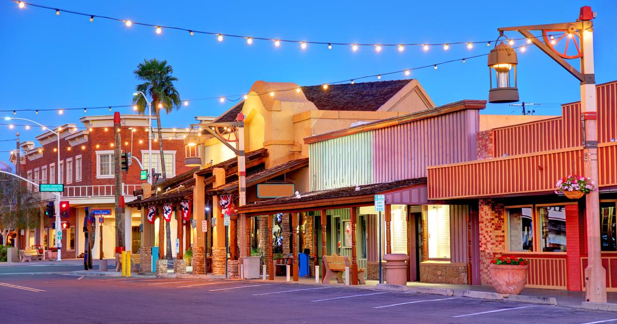 A charming street scene in Old Town Scottsdale is captured at dusk, featuring colorful storefronts with decorative wooden facades. Soft string lights enhance the ambiance, while palm trees provide a touch of greenery against the twilight sky.