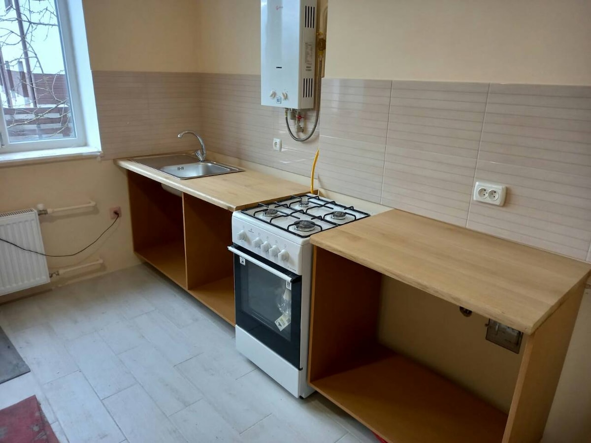 A well-equipped kitchen features a light-colored countertop with open shelving beneath. A stainless steel sink is positioned near a window, while a four-burner gas stove is located on one side. The light-colored tiled walls enhance the space's simplicity.