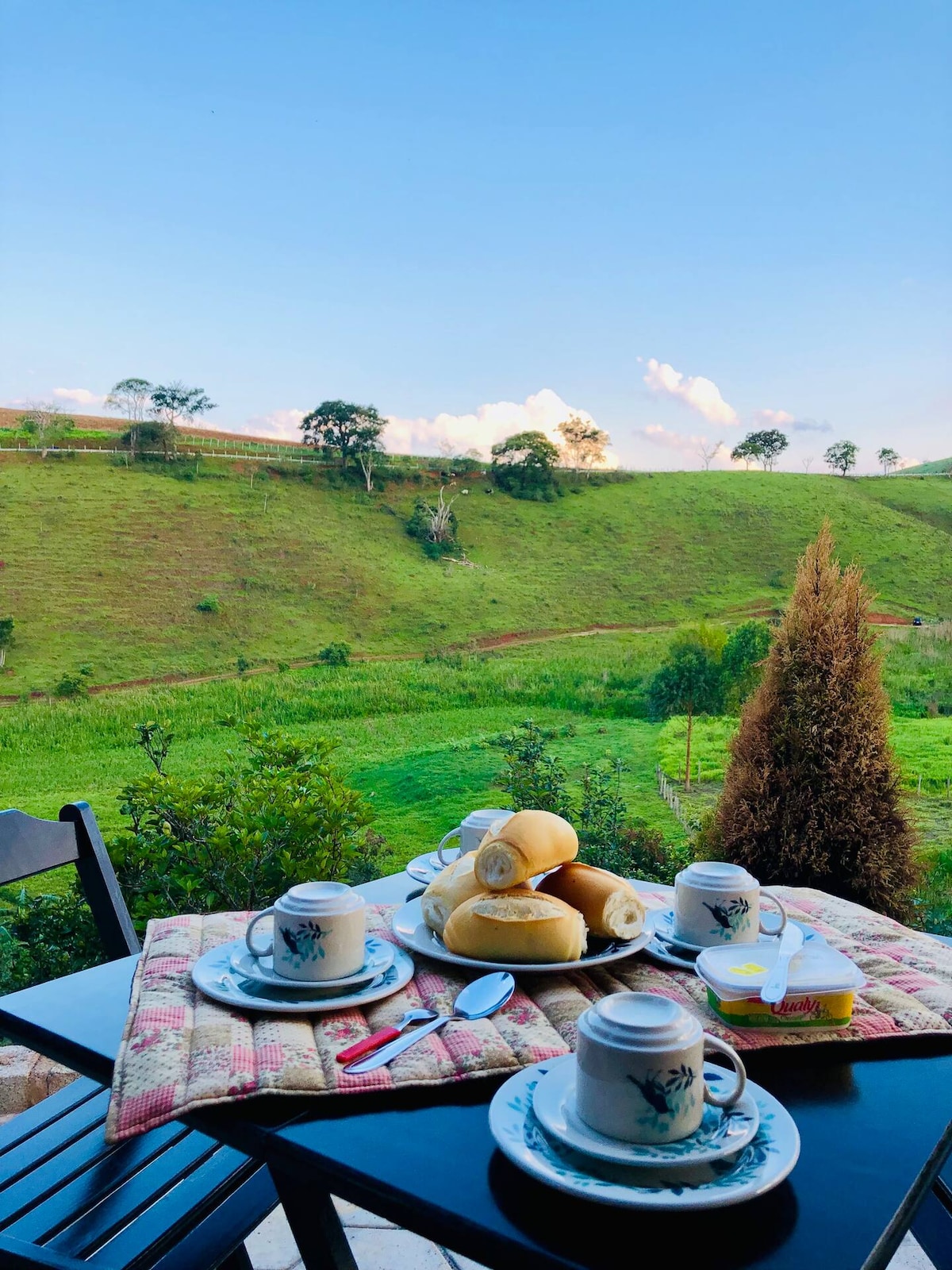 A dining table is set outdoors, featuring plates with fresh rolls, cups, and a small dish of butter. Lush green hills extend in the background under a clear blue sky, providing a serene and picturesque setting.