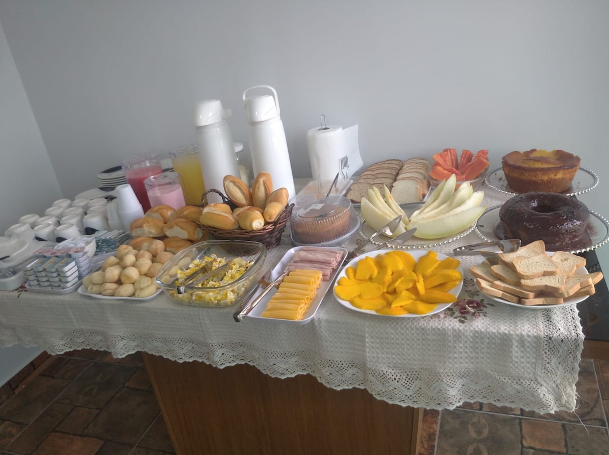 A variety of breakfast items is displayed on a neatly arranged table. Assorted breads, pastries, and fruits are visible, along with desserts and beverages in pitchers. Plates contain sliced cheese and meats, while a lace tablecloth adds a touch of elegance.