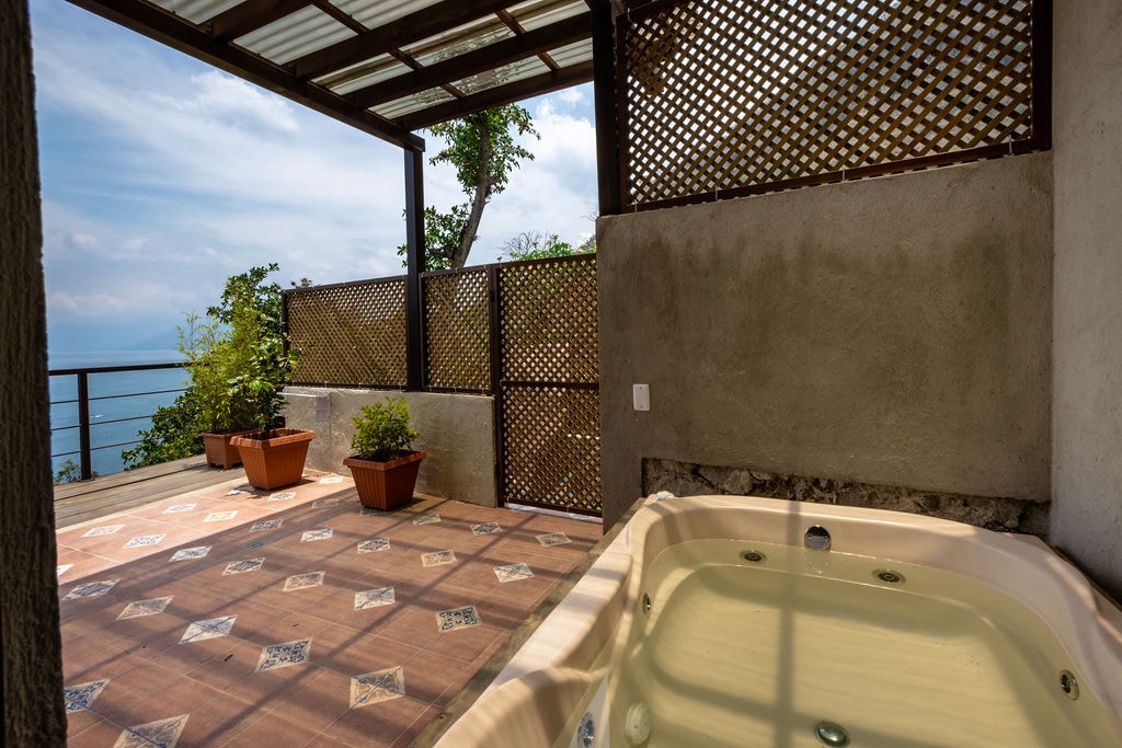 An outdoor area features a jacuzzi positioned alongside potted plants, with views of the lake in the background. The space is sheltered by a lattice roof, allowing natural light to filter through while providing privacy.