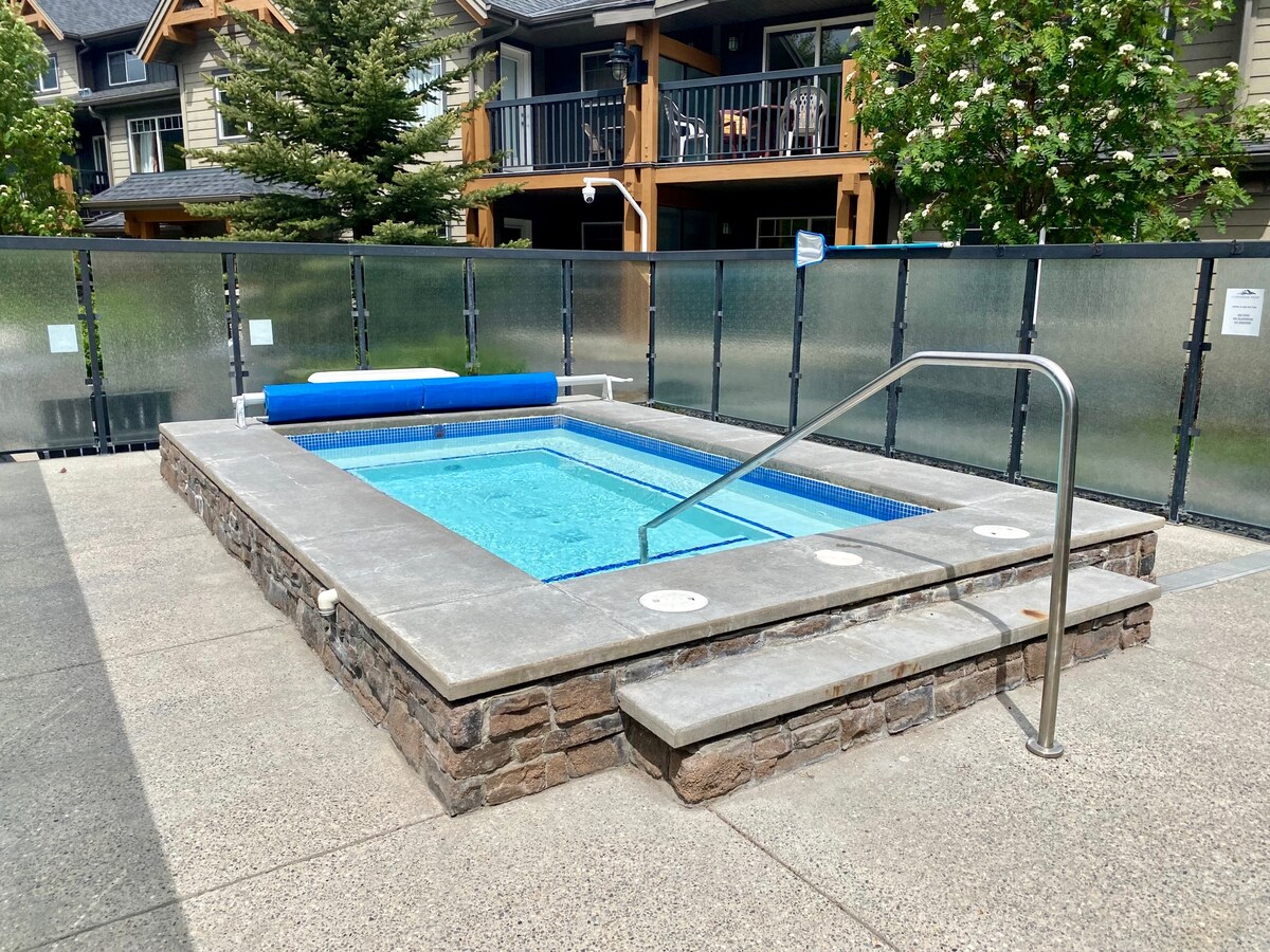 A shared hot tub is situated on a stone patio, featuring a blue cover and stainless steel railing. Surrounding glass panels provide privacy, while trees and townhouses are visible in the background, contributing to a serene environment.