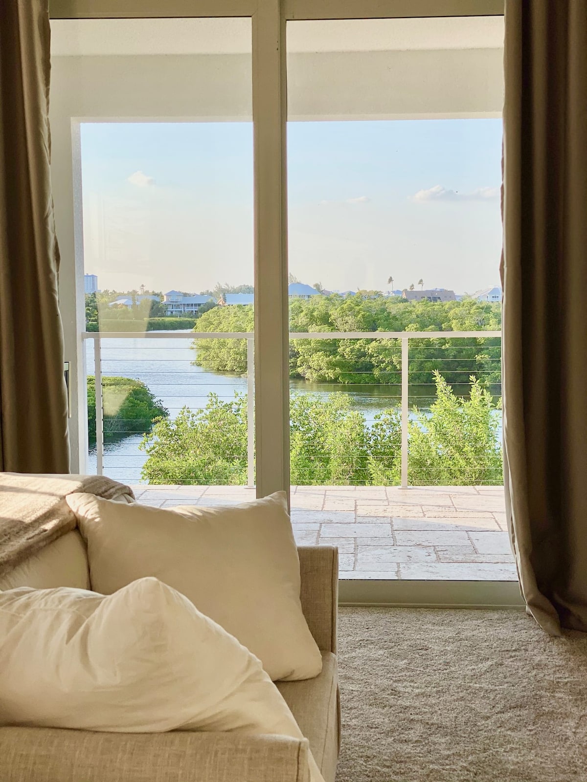 A view of lush greenery and a serene body of water is framed by large glass doors. A spacious, neutral-toned couch with soft pillows sits in the foreground, inviting relaxation. Natural light fills the space, enhancing the tranquil atmosphere.