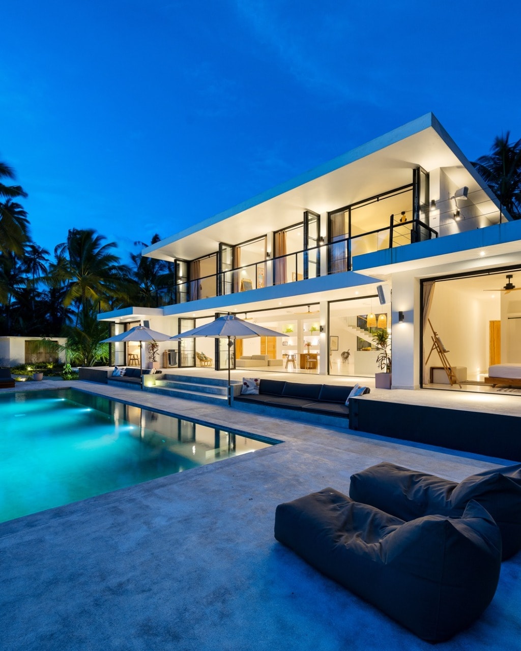 The villa is showcased at dusk, with modern architecture highlighted by large glass doors and balconies. An inviting infinity pool reflects the evening sky, while lounge chairs are arranged beside the water, surrounded by lush tropical vegetation.