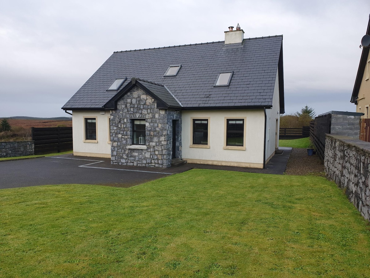 The exterior of the house is depicted, showcasing a two-story structure with a distinctive stone accent and a slate roof. Expansive green lawns surround the property, enhancing the serene setting. Large windows provide ample natural light, and the paved driveway leads to the entrance.