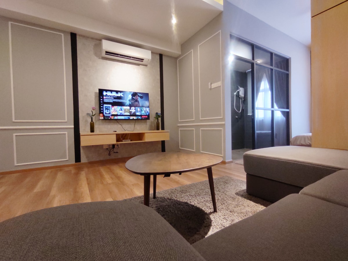 A modern living area features a comfortable sofa and a low coffee table, with a television mounted on the wall. Soft lighting enhances the sleek design, while air conditioning provides comfort. A glass-enclosed bathroom is visible in the background, and a spacious sleeping area is suggested.
