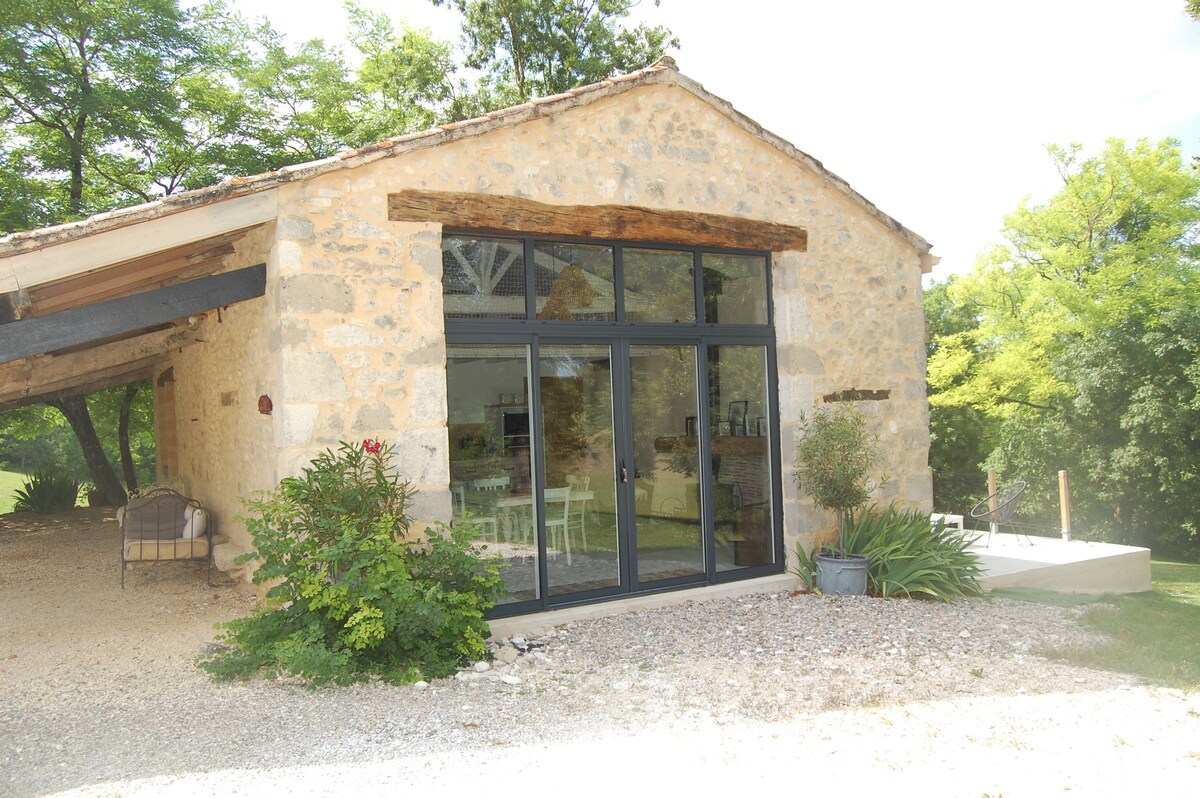 A rustic stone structure features large glass doors that invite natural light into the interior. The surrounding landscape is lush with greenery, framed by trees, creating a serene outdoor atmosphere. A gravel path leads to the entrance, enhancing accessibility.