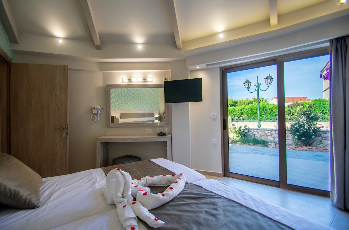 A bright bedroom features a spacious bed with neutral linens. A mirror and flat-screen television are mounted above a streamlined console table. Sliding glass doors open to a patio surrounded by greenery, while soft lighting enhances the room's tranquil ambiance.