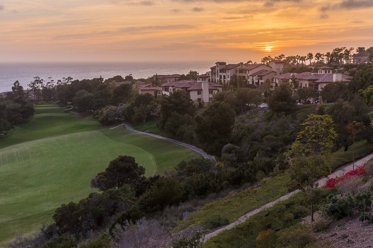 A golf course is set against a stunning sunset, with green fairways leading towards the ocean. The resort buildings are visible in the background, surrounded by lush trees and colorful flowering shrubs that enhance the landscape's natural beauty.