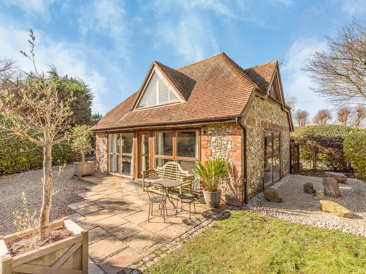 The barn conversion offers a charming exterior with a stone facade and a sloped roof. Outdoor seating is provided on a flagstone patio, surrounded by neatly manicured gardens. Large windows allow ample natural light, while landscaped borders add greenery to the serene setting.