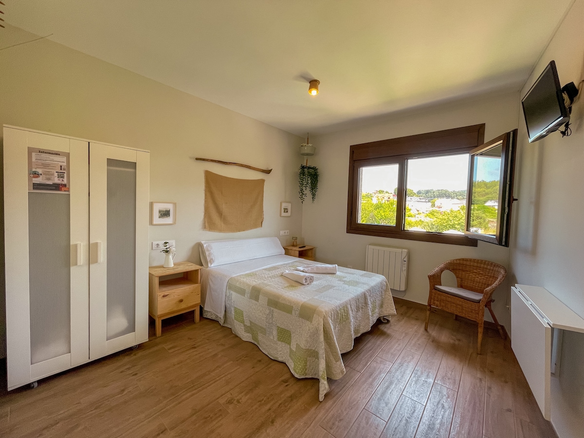 A spacious bedroom features a large bed layered with a light quilt. Natural light pours in through a nearby window with a view. A wicker chair and simple decor complement the wooden furnishings, while a wall-mounted television adds convenience.