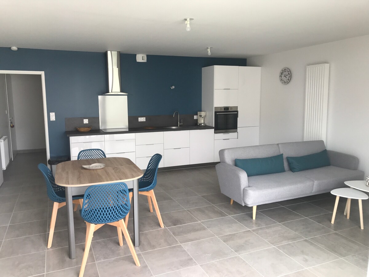 A modern kitchen and living area are presented in a spacious layout. White cabinetry and stainless-steel appliances complement the dark countertop. A round dining table with blue chairs and a gray sofa are visible, set against a blue accent wall. A small side table completes the arrangement.