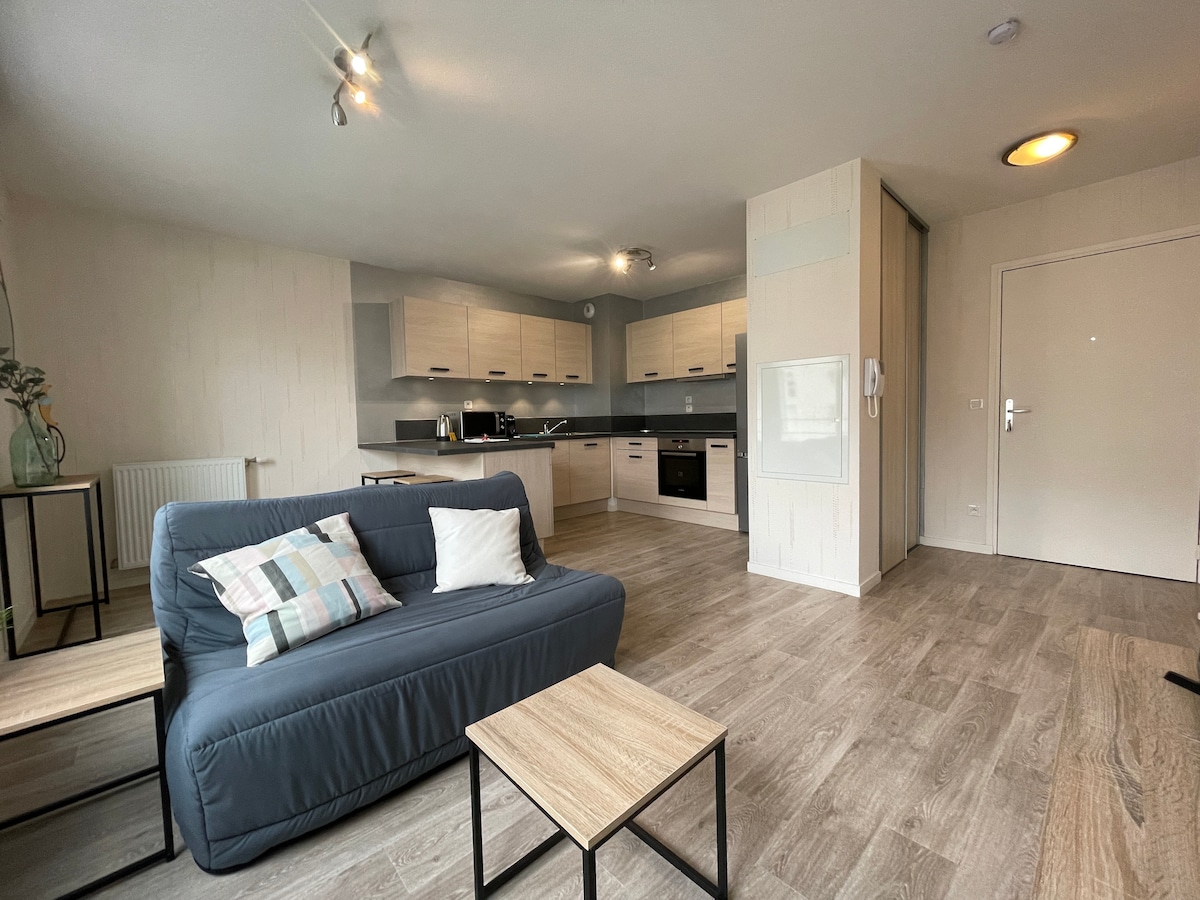 A modern living area features a comfortable gray sofa with decorative cushions, paired with a light wood coffee table. The open kitchen is equipped with contemporary appliances and cabinets, offering ample counter space. Natural light is present, enhancing the inviting atmosphere.
