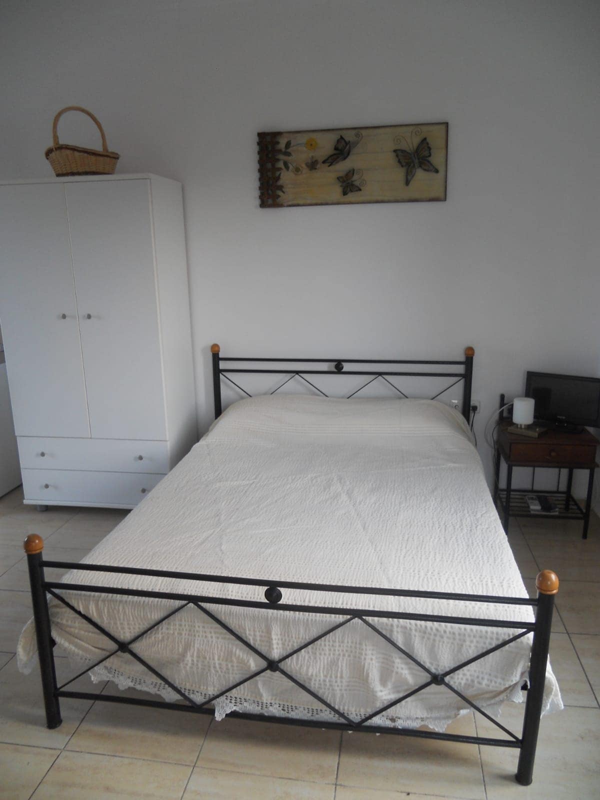 A simple bedroom features a black metal bed with a light-colored blanket. A white wardrobe is positioned beside the bed, while a small table with a lamp and a television are also present. A wall decoration with floral motifs adds a touch of character.