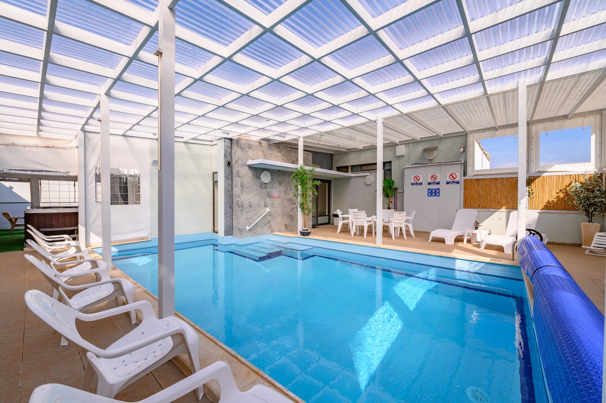 A well-lit indoor pool area features a heated pool with a seating area nearby. Several lounge chairs are positioned around the pool, and natural light filters through a spacious, translucent roof. Walls are accented with neutral tones and greenery, contributing to a relaxing environment.