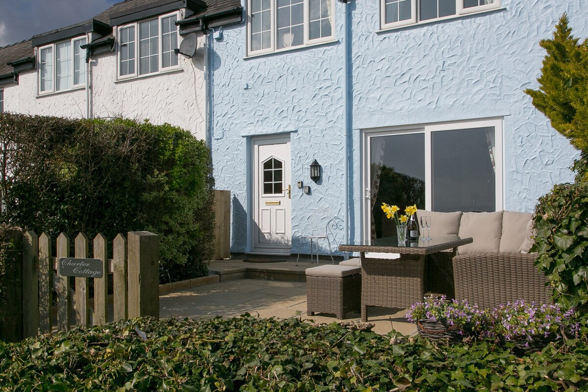 The exterior view of the cottage presents a charming blue facade set against greenery. A patio area features comfortable seating with a table, and bright yellow flowers add a touch of color. Large glass doors provide a view into the inviting interior.