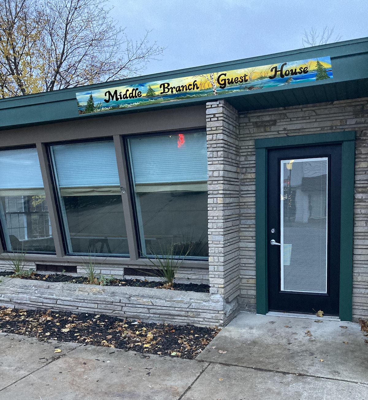 The exterior of the Middle Branch Guest House is prominently displayed with a colorful sign above the entrance. Large windows provide natural light, framed by neatly arranged plantings and stonework. The front door features a glass panel, enhancing accessibility.