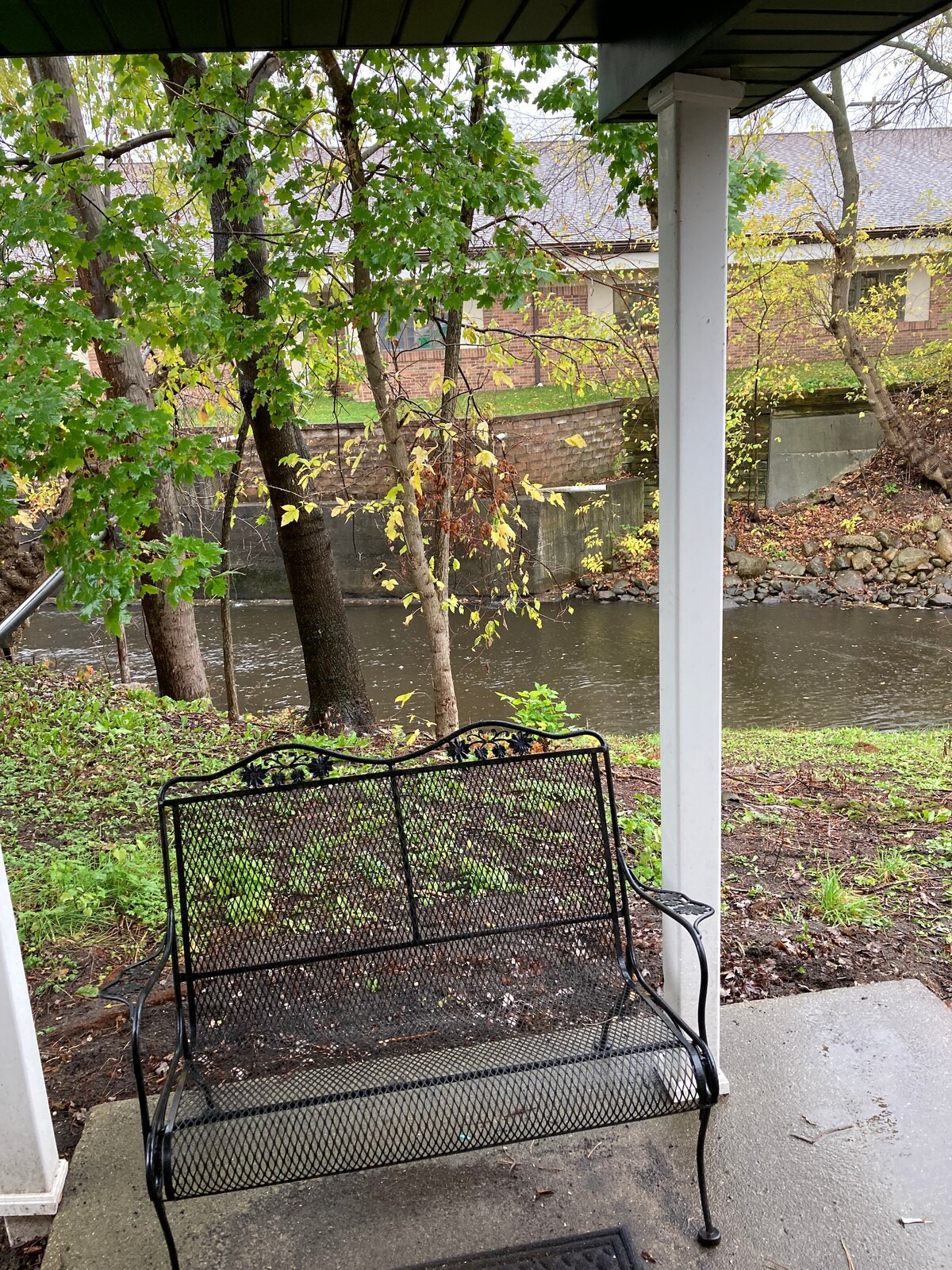 A black metal bench is positioned under a covered porch, facing a flowing river. Trees, adorned with autumn leaves, frame the scene, and the water gently reflects the surrounding nature. The ground is slightly damp, indicating recent rainfall.