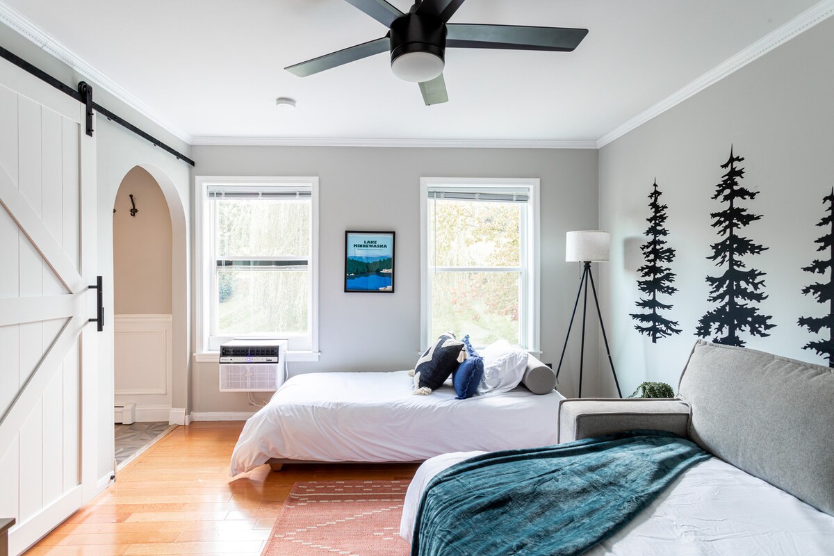 A serene bedroom presents two spacious beds, each with clean bedding. A cozy gray couch is positioned nearby. Natural light fills the room through multiple windows, revealing a tranquil view of trees outside. A fan overhead provides a sense of openness.