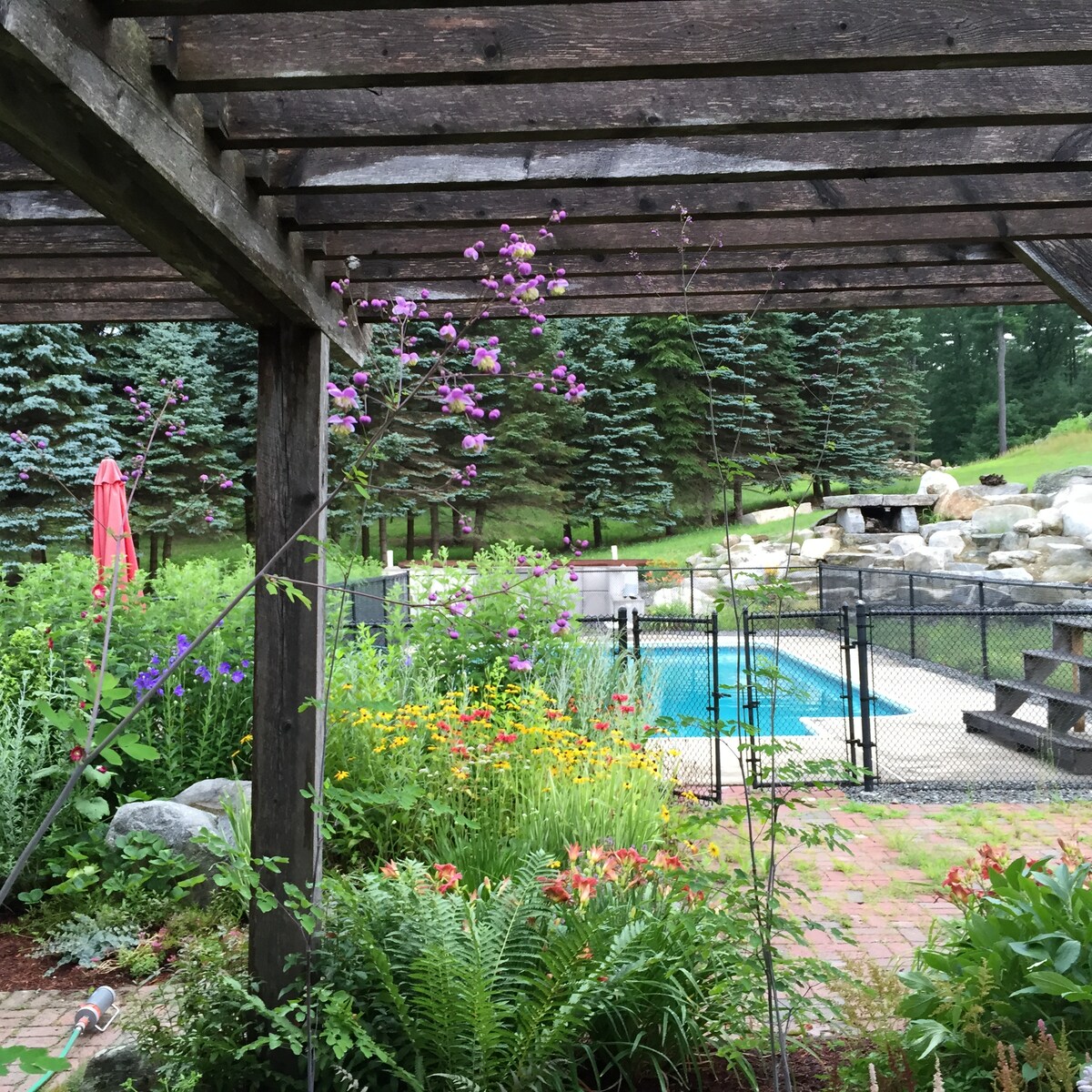 A tranquil outdoor area is framed by wooden beams, showcasing a vibrant garden filled with colorful flowers. A fenced pool is visible in the background, surrounded by lush greenery and stone features. A patio area is indicated by stone steps leading away from the garden.