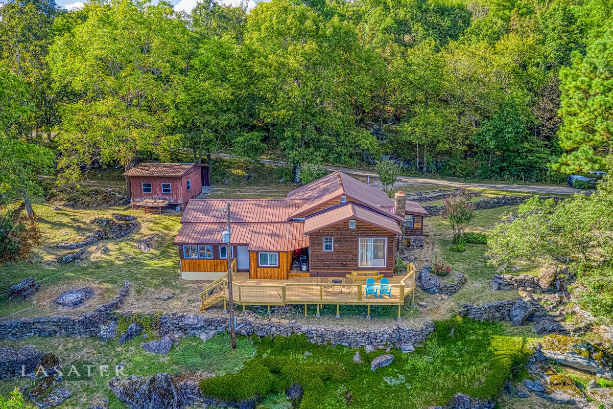 An aerial view showcases a rustic log cabin, surrounded by lush greenery and large boulders. The cabin features a deck with two blue chairs, inviting outdoor relaxation. Nearby, a smaller structure adds to the property’s charm, all enveloped by a serene natural landscape.
