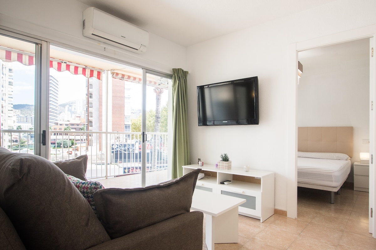 A bright living area features a comfortable sofa facing a wall-mounted television. Large glass doors open to a terrace, allowing natural light to fill the space. A doorway leads to the bedroom, which includes a spacious bed and light-colored furnishings.