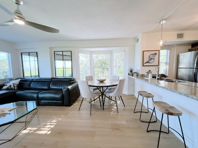 A modern open-concept living area features a black sectional sofa and a round dining table surrounded by white chairs. Natural light floods the space through large windows, which frame views of lush greenery. A sleek kitchen with granite countertops is visible in the background.