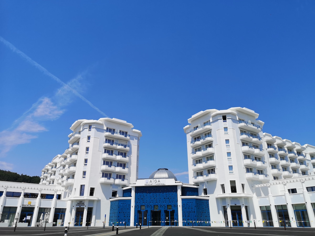 A modern building is showcased with a white facade and distinctive blue accents. Multiple balconies can be observed on the upper floors, while a central dome adds architectural interest to the entrance. The clear blue sky provides a bright background.