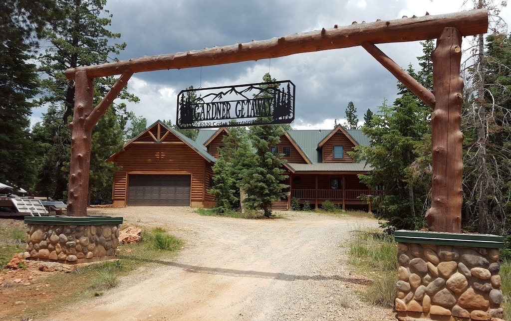A large log archway marks the entrance to the mountain home, featuring a decorative sign with 'Gardner Getaway.' The surrounding area is characterized by tall trees and a gravel driveway leading to a spacious wooden house with a green metal roof.